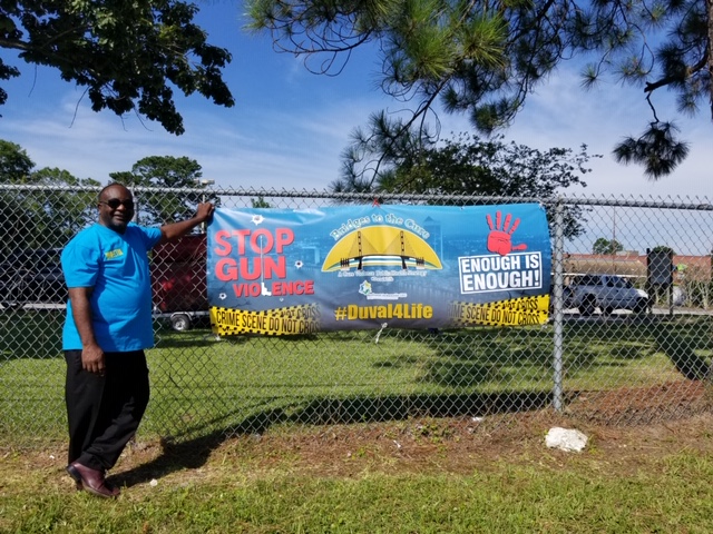Paul Tutwiler: Man in sunglasses, blue T-shirt, black pants, brown shoes stands to left of poster on fence that says stop the gun violence Duval4life enough is enough