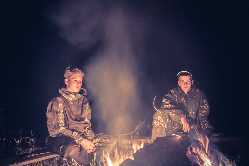 Boy Scouts: 2 serious boys in Scout uniforms sit before fire in darkness