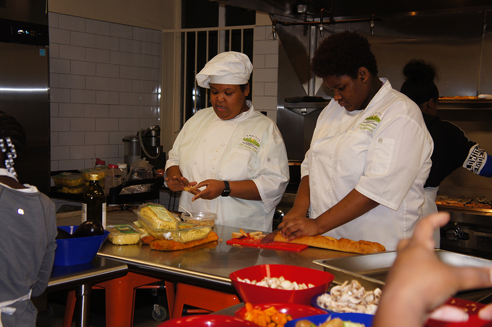 cooking: 2 women in white cooking uniforms work with food at steel counter