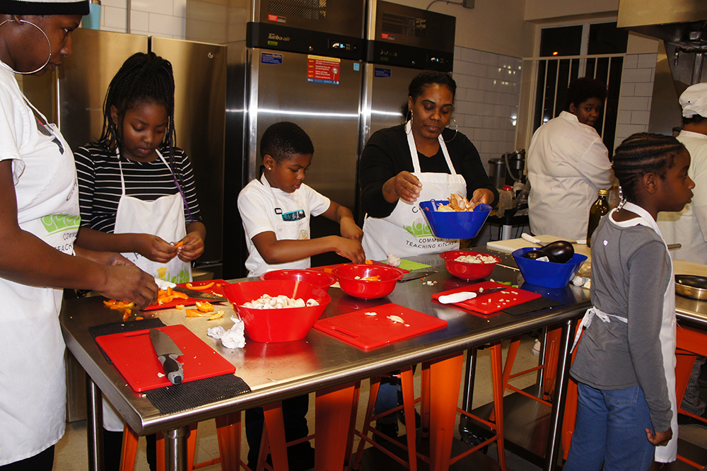 cooking: Kids and adults in white aprons cook at steel counter with red chopping boards and bowls.