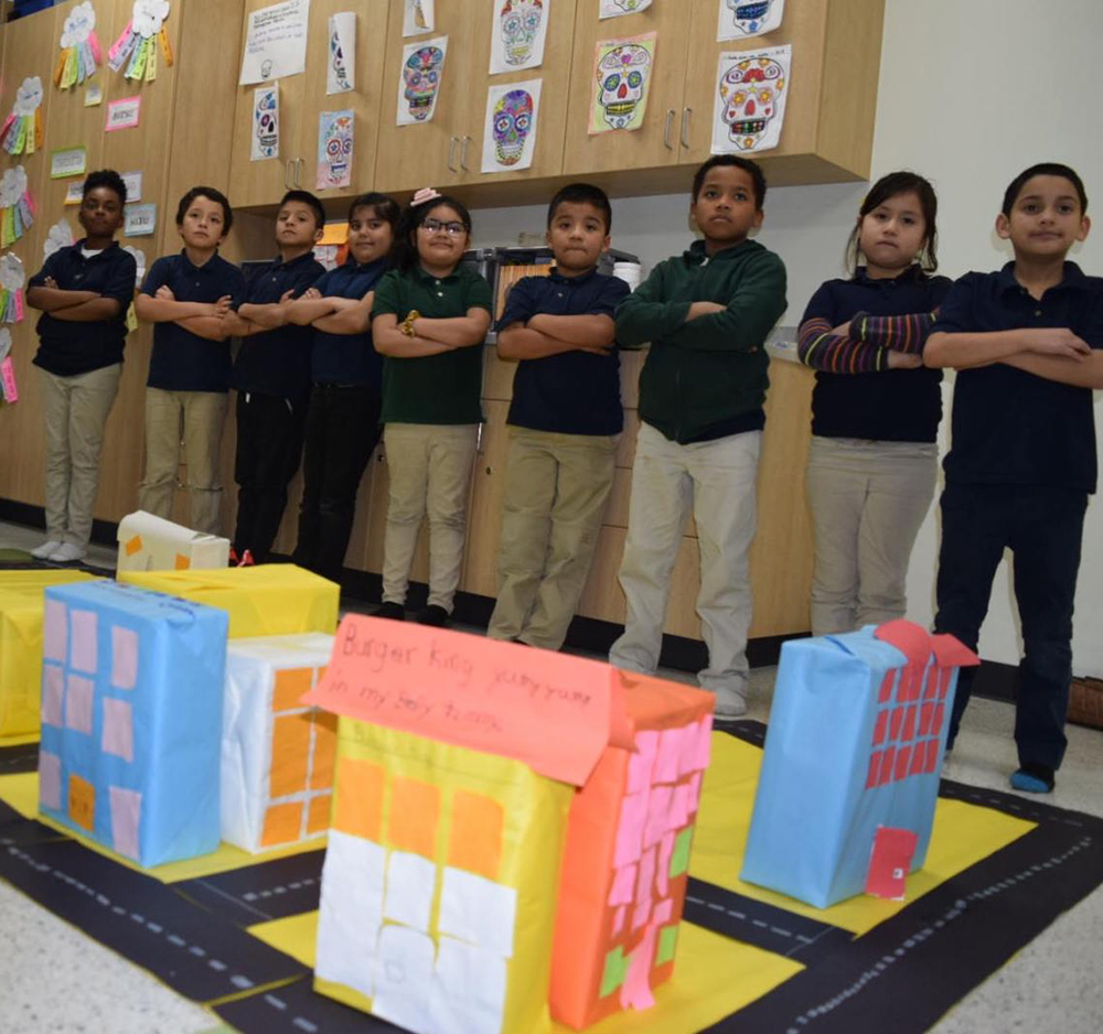 Greenwood: Children’s town built from construction paper; line of kids with folded arms stand behind it