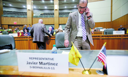 New Mexico: Man on phone at desk in large chamber with other people in background. Desk holds name card, 2 small flags.