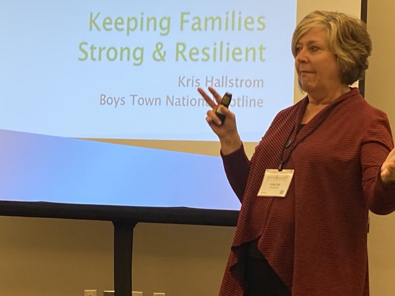 suicide: Woman with short, wavy hair, wearing ID badge on lanyard and orange outfit, gestures in front of screen that says keeping families strong and resilient, Kris Hallstrom.