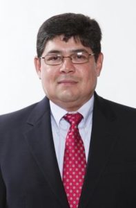 LGBTQ: Sam Terrazas (headshot), professor at Facundo Valdez School of Social Work, man with short dark hair, glasses, wearing dark suit, white shirt, red patterned tie