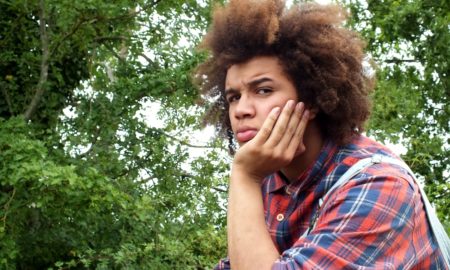 trauma: teenage boy with afro hair, looking angry, resting his face in his hand, in the countryside , wearing a red check shirt