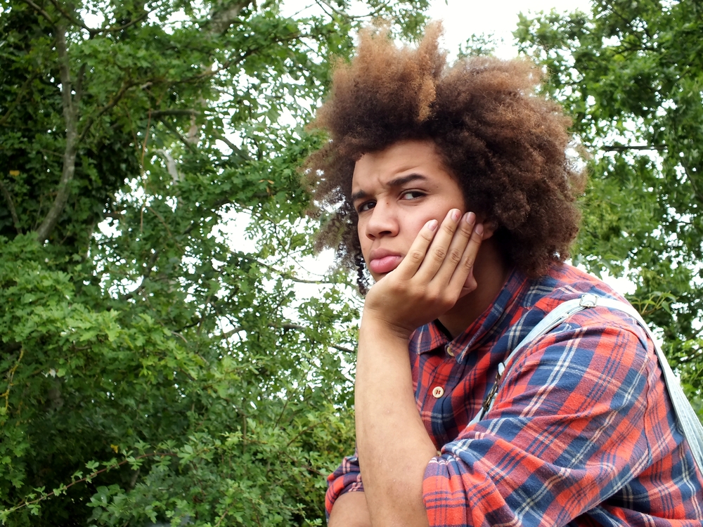 trauma: teenage boy with afro hair, looking angry, resting his face in his hand, in the countryside , wearing a red check shirt