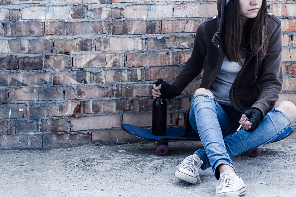 homeless: Young woman in hood is sitting on skateboard and holding smoldering cigarette and bottle of beer.