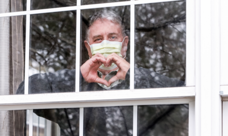 COVID-19 Community Healthcare and Education grants; man wearing medical mask in window making heart shape with hands