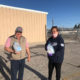 Navajo: Man and woman stand outside holding jugs of water. He’s smiling, wearing cap with U.S. flag on it. She’s wearing mask.