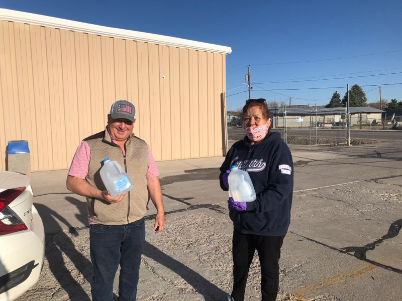 Navajo: Man and woman stand outside holding jugs of water. He’s smiling, wearing cap with U.S. flag on it. She’s wearing mask.