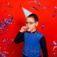 autism: A boy in a festive cap blows into a festive pipe and stands in the rain of blue and silver confetti on a red background.