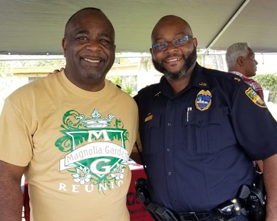 2 smiling men, one in T-shirt, the other in blue police or security officer uniform, stand under tent outside.