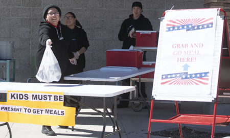 Gallup: Woman in cap, jacket holds out full white plastic bag at table next to sign that says grab and go meal here