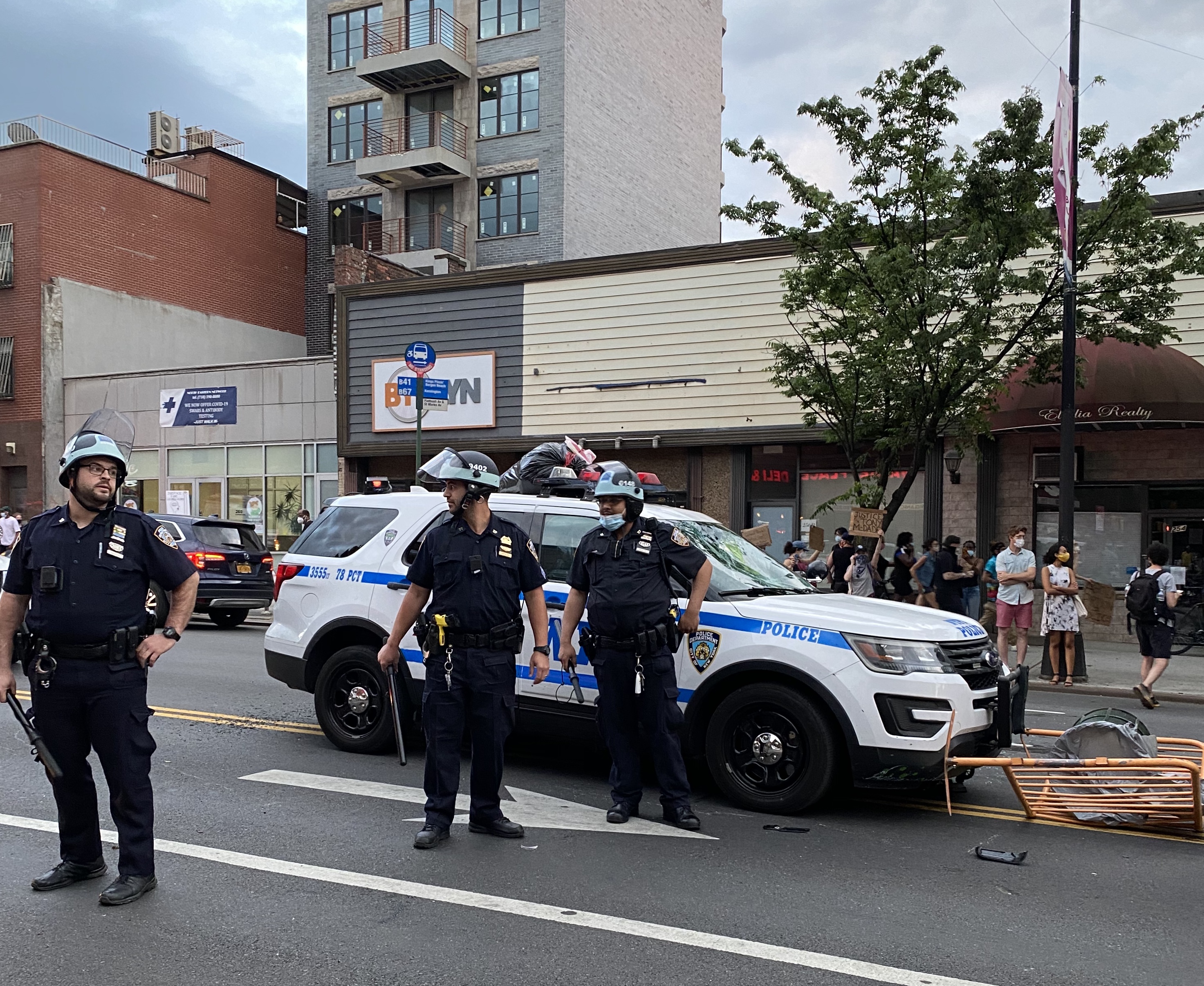 3 New York Police officers stand near squad car in middle of street.