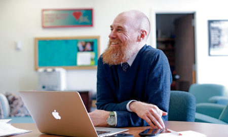 extended foster care: Beaming man with red beard, blue sweater, shirt, tie sits at laptop at desk
