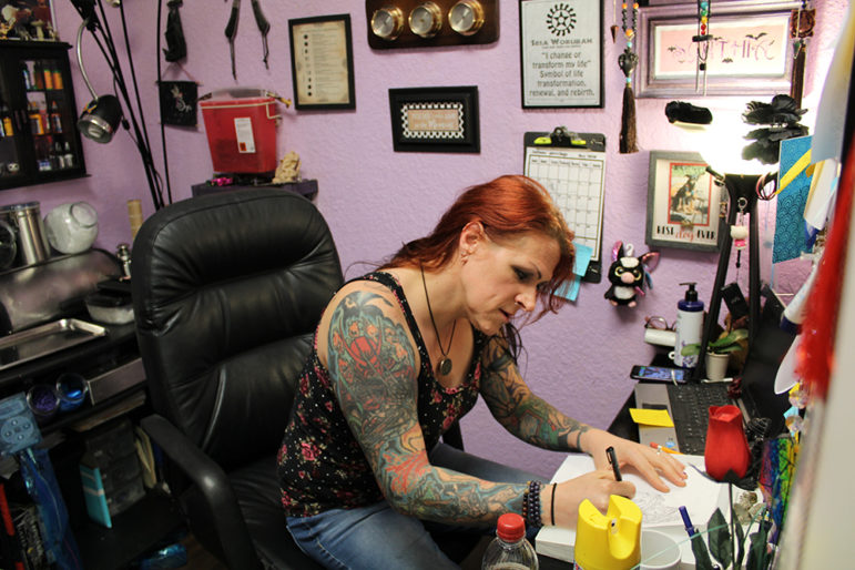 LGBT: Woman with long red hair, 2 tattoo sleeves in flowered tank top works at desk