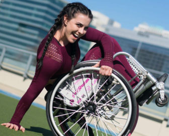 money: smiling woman with long brown hair in braid, maroon sweater tilts back wheelchair on grass