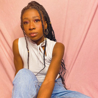disabilities: Woman with long black braids, earrings, nose ring wearing white sleeveless top, faded jeans poses against apricot background