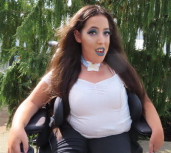 frontliners: Woman with long brown hair, blue makeup, white top in wheelchair outside