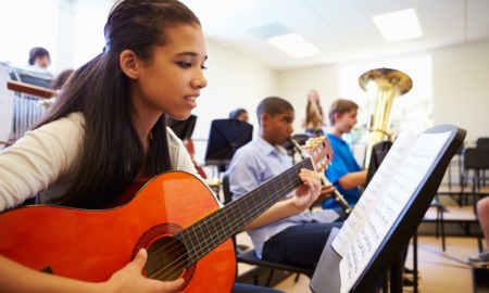 Youth music education program grants: young ethnic female playing guitar in room filled with students playing instruments