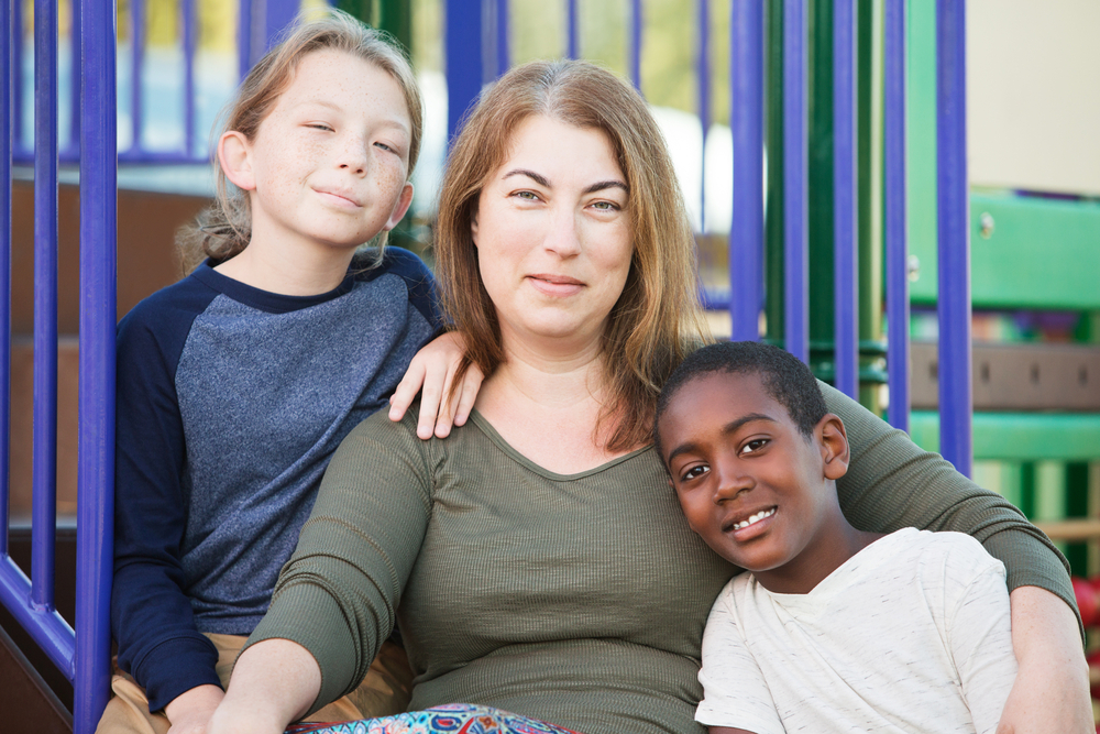 caregivers: White woman sitting in playground with 2 boys, one black and one white