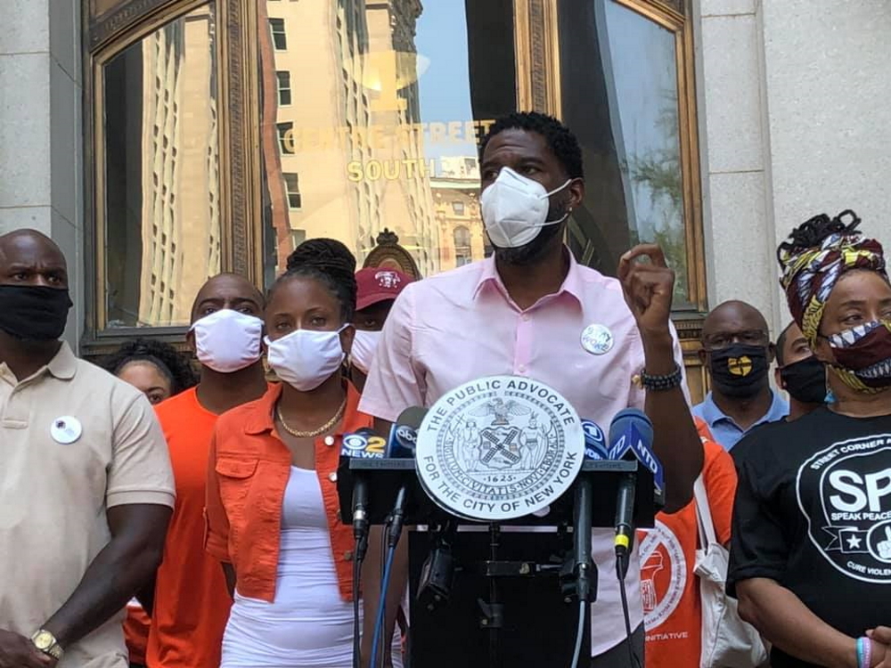 gun violence: Man in short-sleeved pink shirt, mask speaks at outdoor podium surrounded by crowd wearing masks