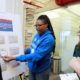 social media: Woman with braids, bright blue hoodie puts cards in envelopes on poster board on stand as several people stand around watching.