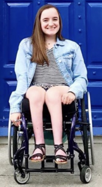 disabilities: smiling woman with long brown hair, striped top, light blue jacket in wheelchair outside