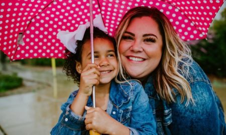 foster care: smiling woman, child in blue denim jackets holding pink polka-dotted umbrella above them