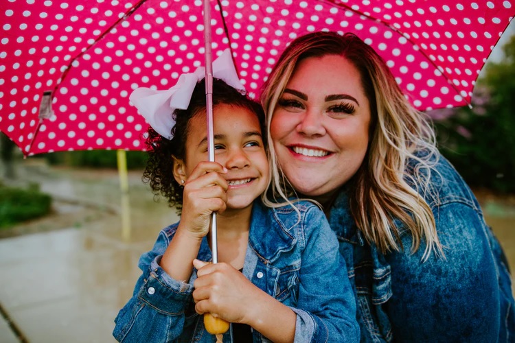 foster care: smiling woman, child in blue denim jackets holding pink polka-dotted umbrella above them