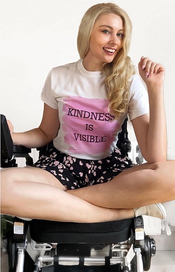exercise: smiling woman with long blonde hair sits cross-legged in wheelchair, wearing T-shirt that says kindness is visible