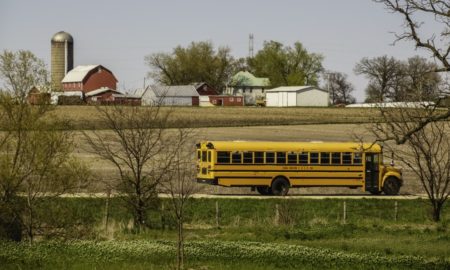 Rural Oklahoma K-12 classroom enrichment and support grants; A public school bus passes by a large farmstead along a rural road on a spring day, with the edge of a county forest preserve in the foreground.