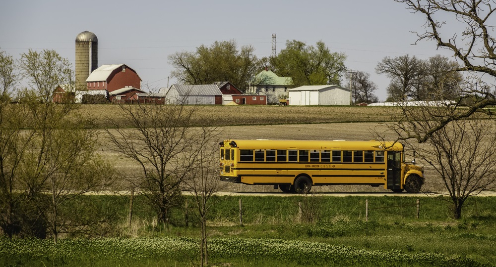 A public school bus passes by a large farmstead along a rural road on a spring day, with the edge of a county forest preserve in the foreground.