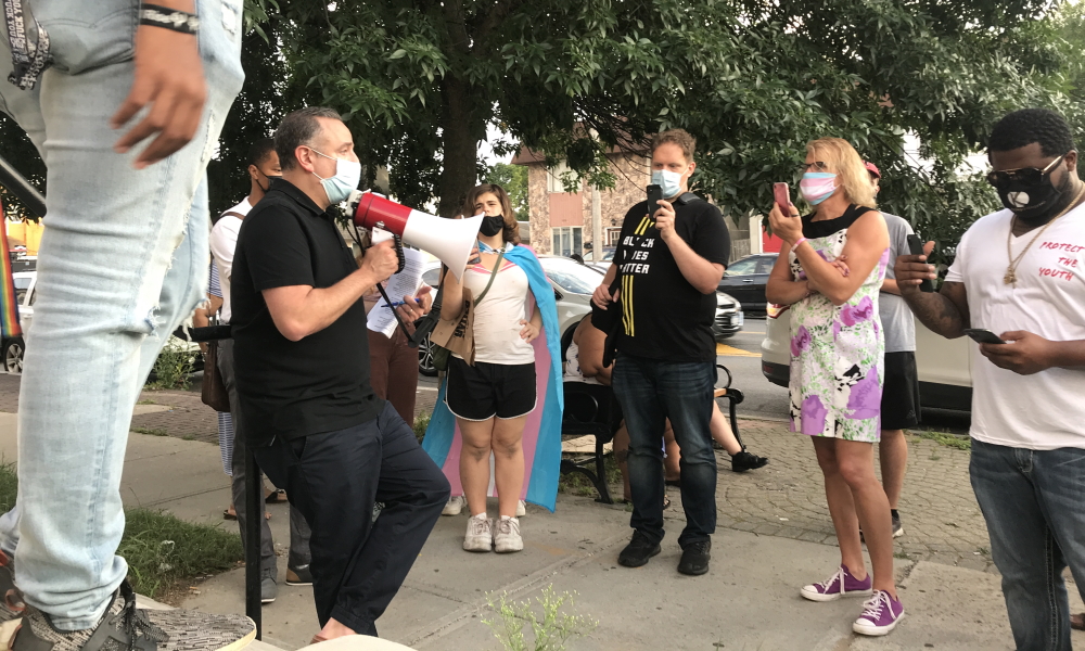 Schenectady: Man in black with megaphone wearing mask talks to small group wearing masks, holding up phones. One woman is wearing superhero-style cape.