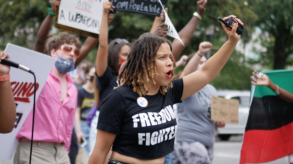 defunding: Woman in protest with short coppery curls, wearing short black T-shirt with message freedom fighter, raises hand holding 2-way radio