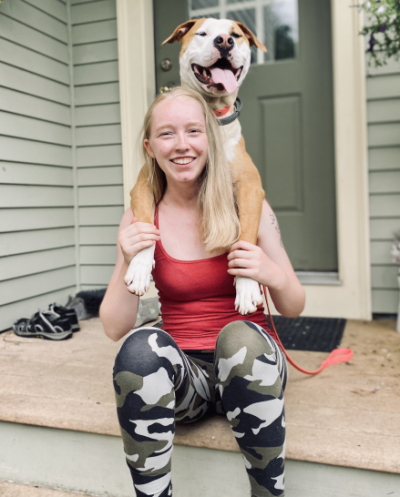 education with disabilities & COVID-19: smiling woman with long blonde hair wearing red tank top and camouflage leggings sits on porch holding large dog’s paws over her shoulders