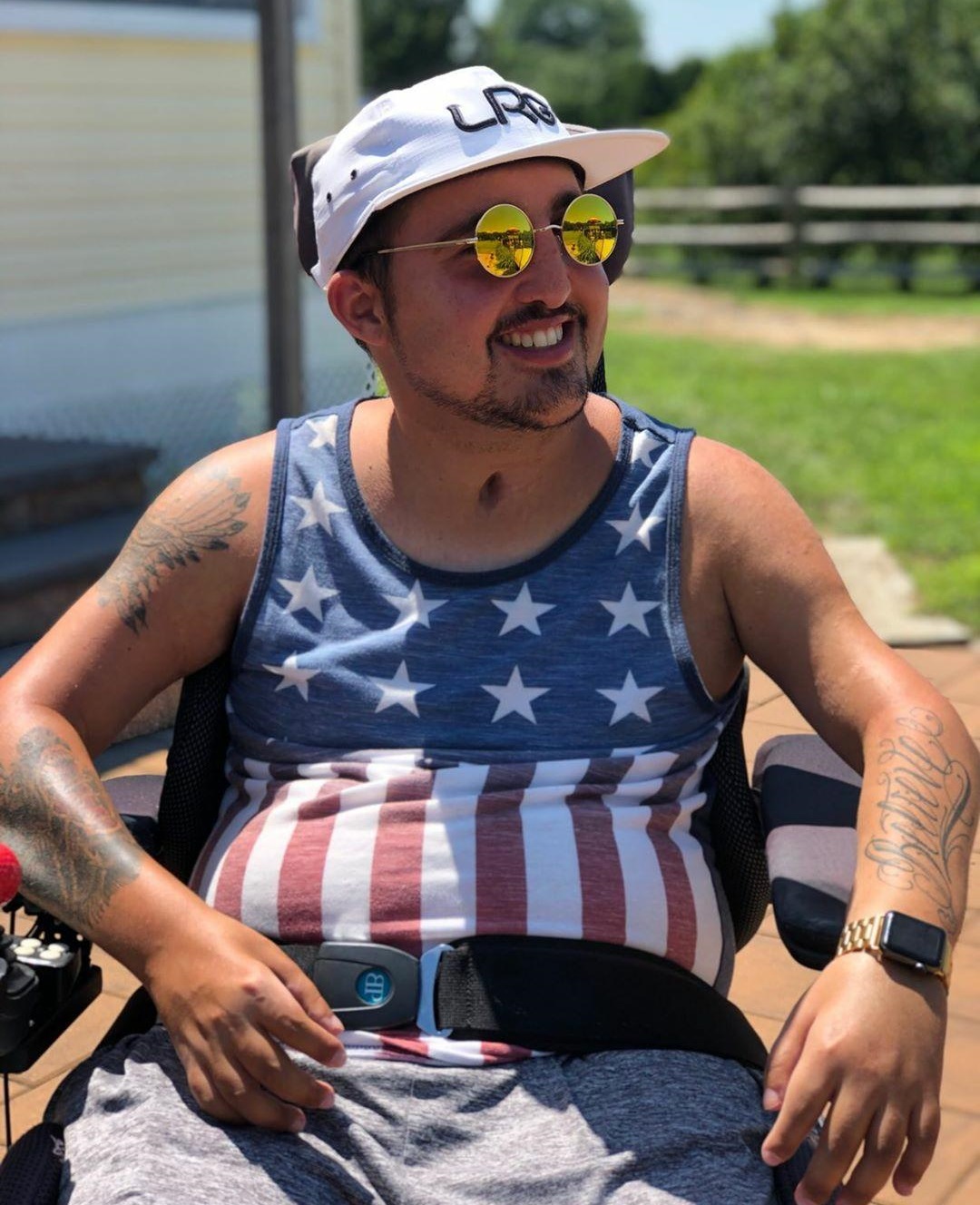 voting: smiling, tan, tattooed man wearing sunglasses, ballcap, flag tank top in wheelchair