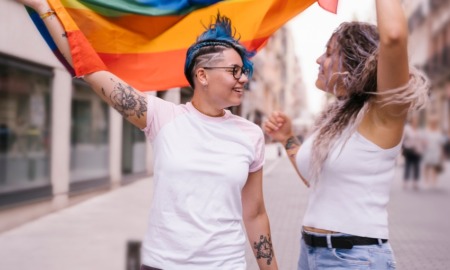 Breaking Barriers to Quality Mental Health Care for LGBTQ Youth report; Lesbian couple with playful attitude showing proudly a pride lgbt flag.