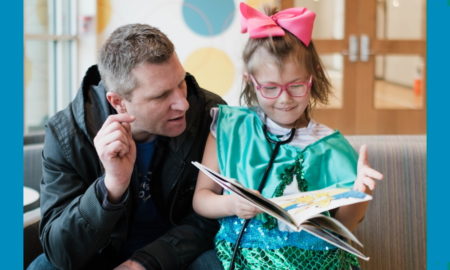 Positive Tomorrows: Beaming little girl holds open book as man on couch looks at it.