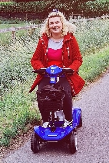 accessibility: smiling woman in red coat sitting on scooter next to field