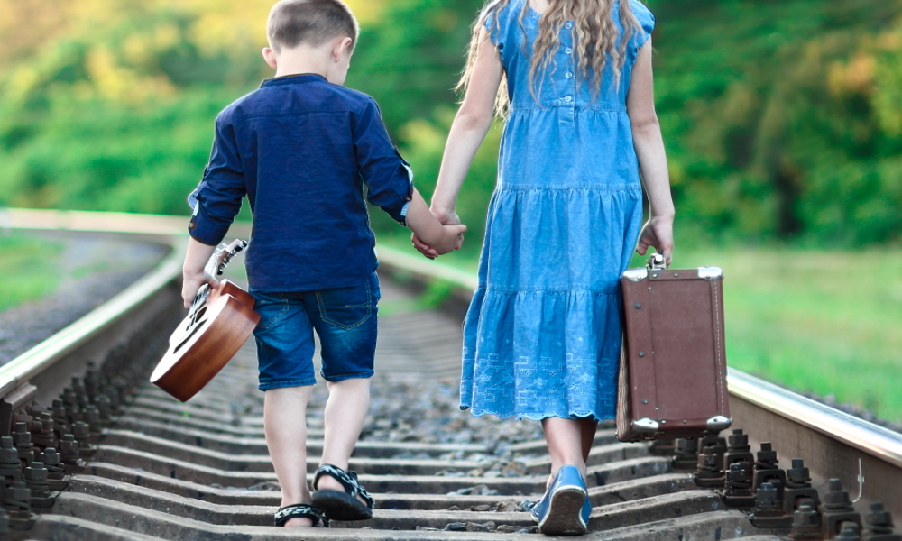  Siblings Foster Care: Girl carrying suitcase and younger boy carrying guitar walk on railroad tracks holding hands, seen from back