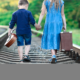 Girl carrying suitcase and younger boy carrying guitar walk on railroad tracks holding hands, seen from back