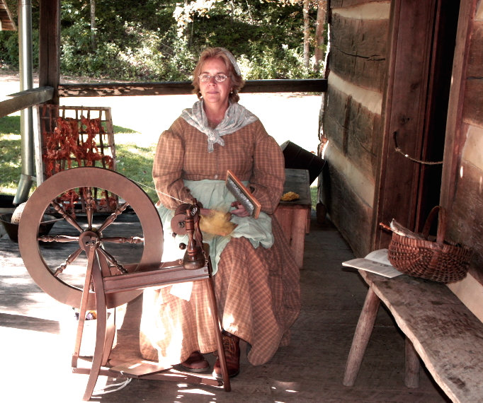 Woman in long dress, kerchief sits before spinning wheel on wooden porch