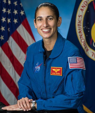 astronaut: Smiling woman with dark hair in blue jumpsuit with patches in front of U.S. flag.