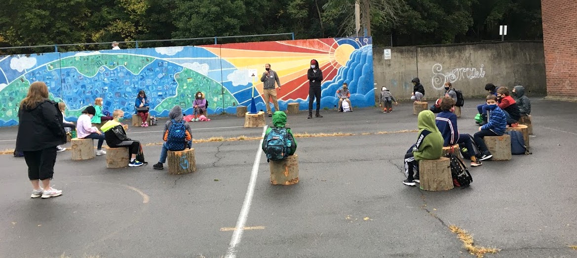 Children in jackets sit on tree stumps outside near colorful mural.