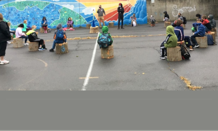 Children in jackets sit on tree stumps outside near colorful mural.