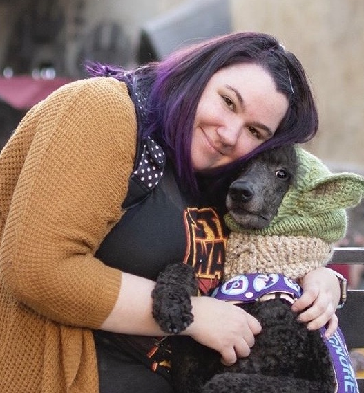 protests: smiling woman with purple hair, orange cardigan bent over dog wearing knit hat