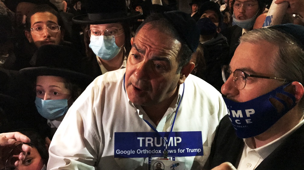 Borough Park: Man in middle of crowd of masked males wearing black hats has Trump sticker on shirt