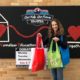 Montana: Smiling woman holds tote bags of different colors in front of sign on brick wall