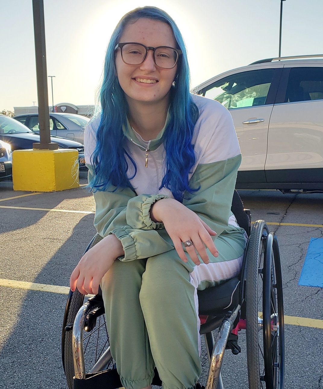 health insurance: smiling woman with blue hair, glasses, wearing white and khaki sports pants, top in wheelchair in parking lot